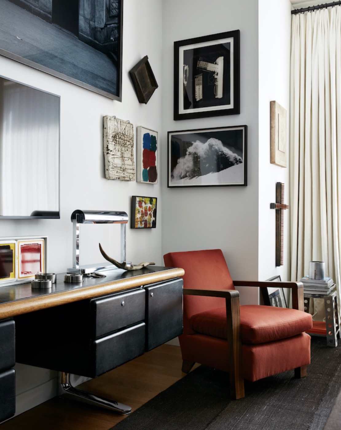 A corner desk with a red-orange lounge chair beneath a group of small framed works