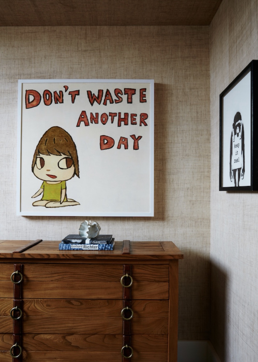 A wooden chest of drawers with brass ring pulls beneath a framed text artwork